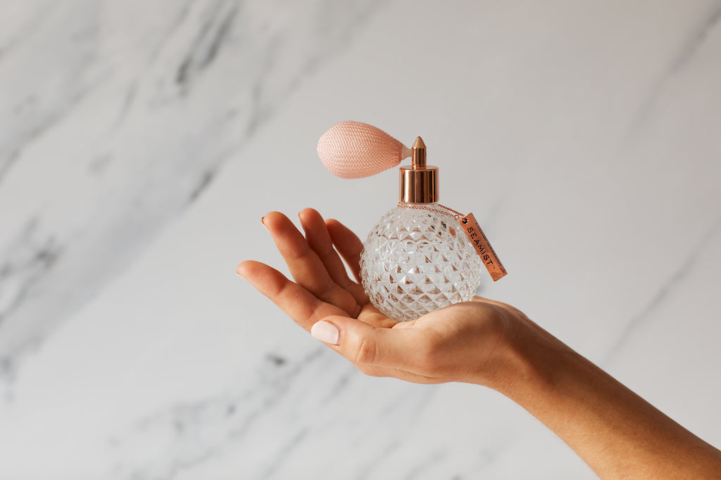Seamist Liquid Garnish in eleganter Kristallflasche mit roséfarbenem Zerstäuber, präsentiert auf einer Hand vor Marmorhintergrund.