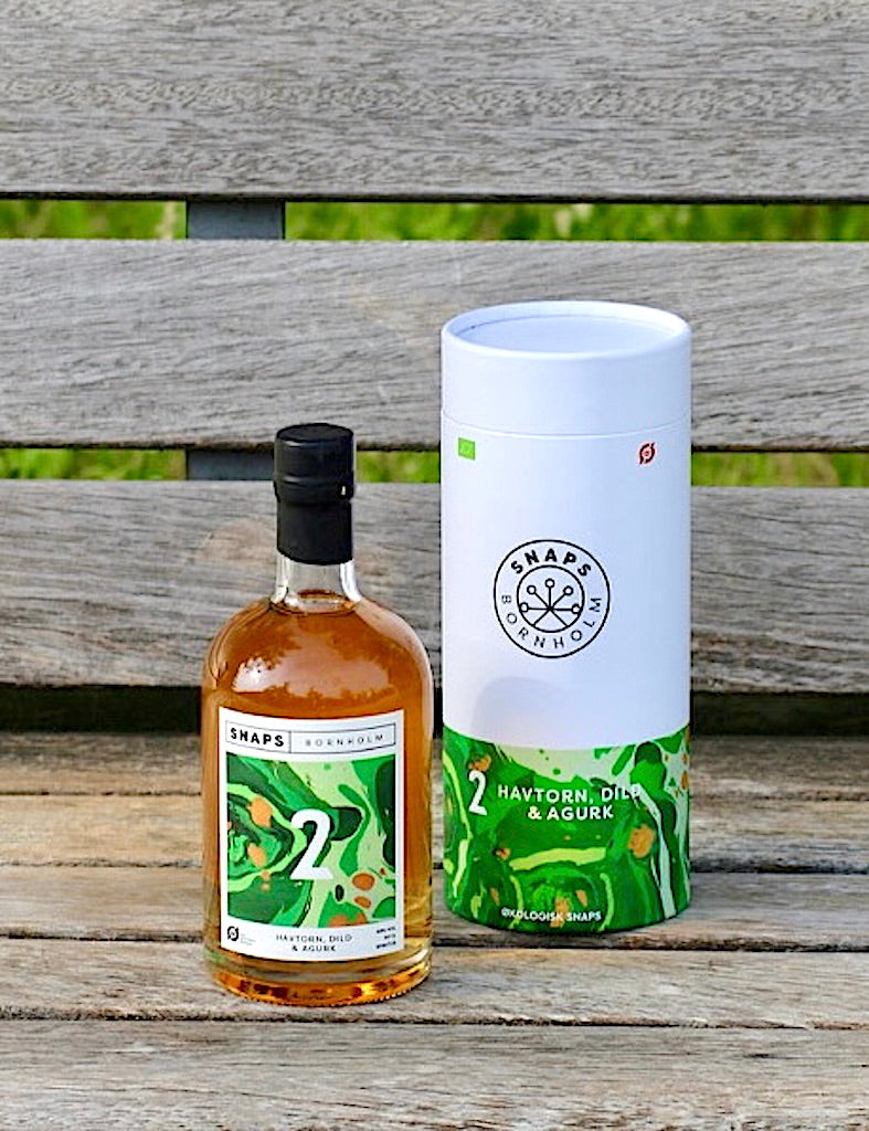 Eine Flasche Bio-Schnaps von Bornholm, Sanddorn, Dill und Gurke, neben der passenden Geschenkbox, stehend auf einer Holzbank.