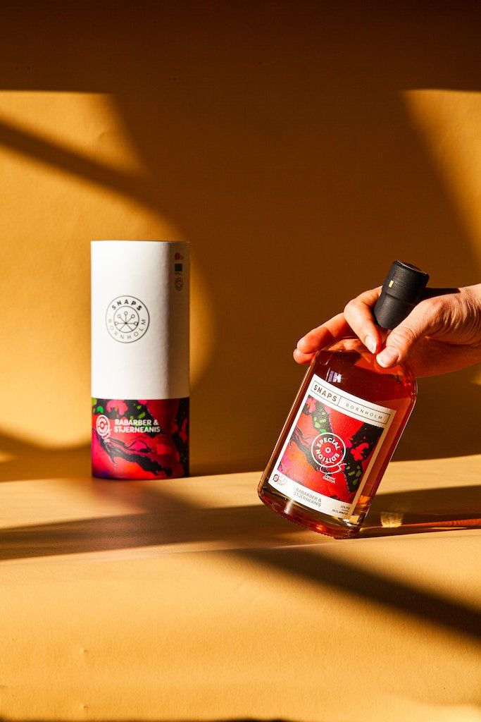 Bio-Schnaps Bornholm Rhabarber & Sternanis in Geschenkverpackung, Flasche mit schwarzem Verschluss in Hand, goldenes Hintergrundlicht.