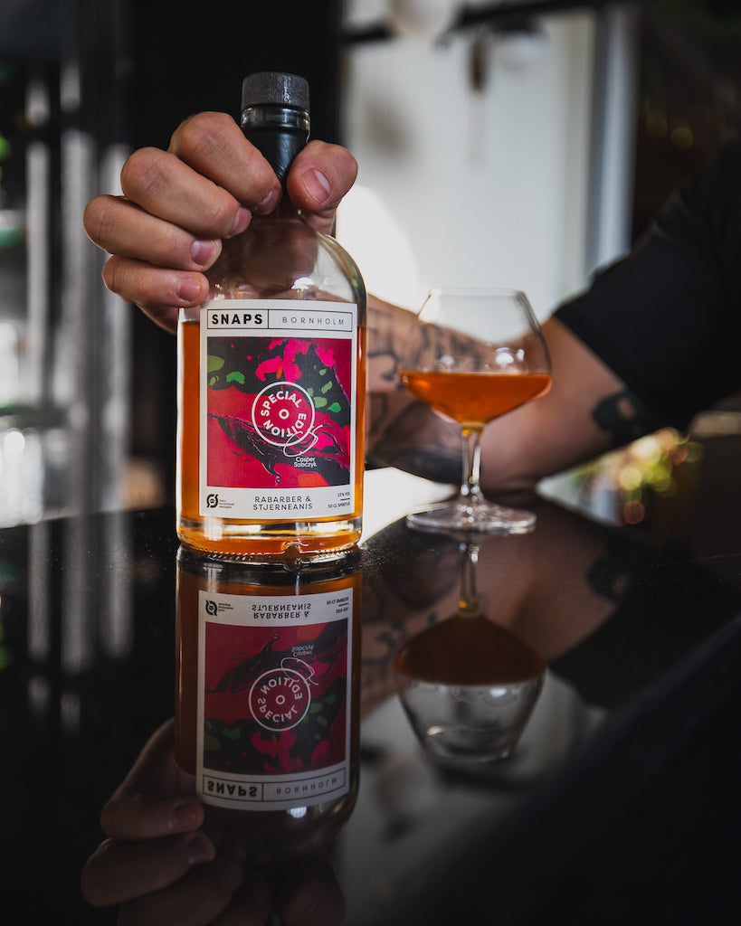 Hand hält eine Flasche Bio-Schnaps Bornholm Rhabarber & Sternanis, Spiegelung auf schwarzem Tisch, daneben ein gefülltes Glas.