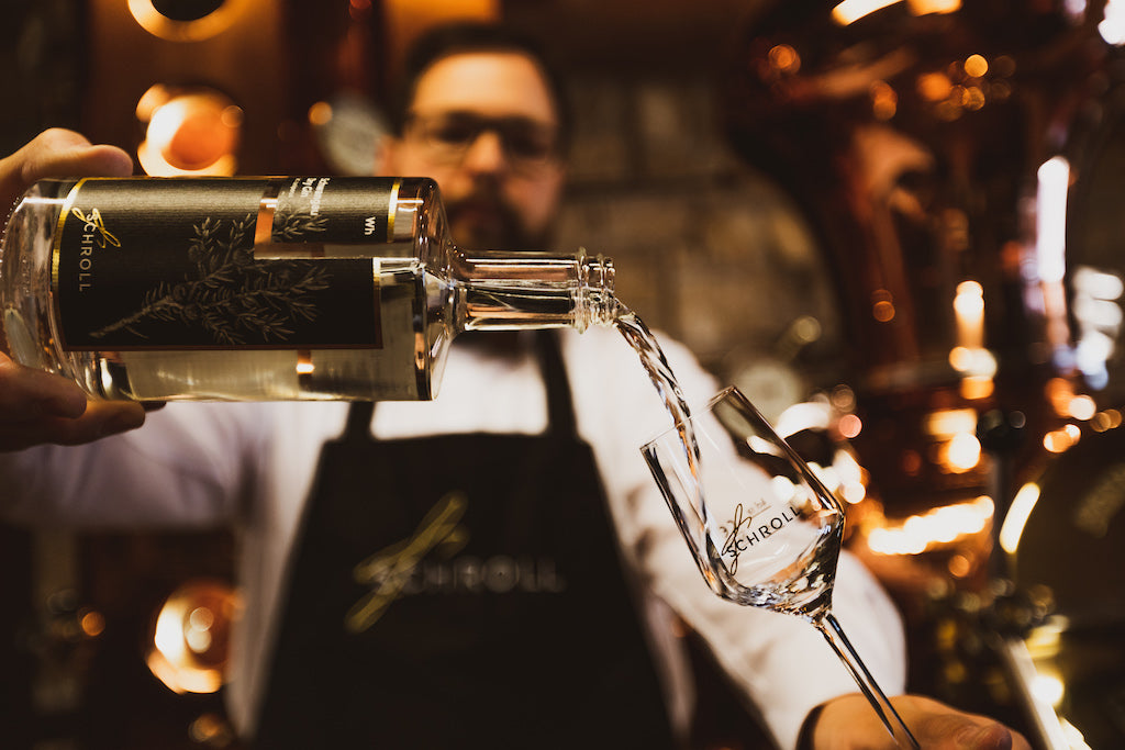 Schwangau Dry Gin wird aus einer Flasche in ein Glas der Brennerei Schroll eingeschenkt, im Hintergrund eine kupferne Destille.