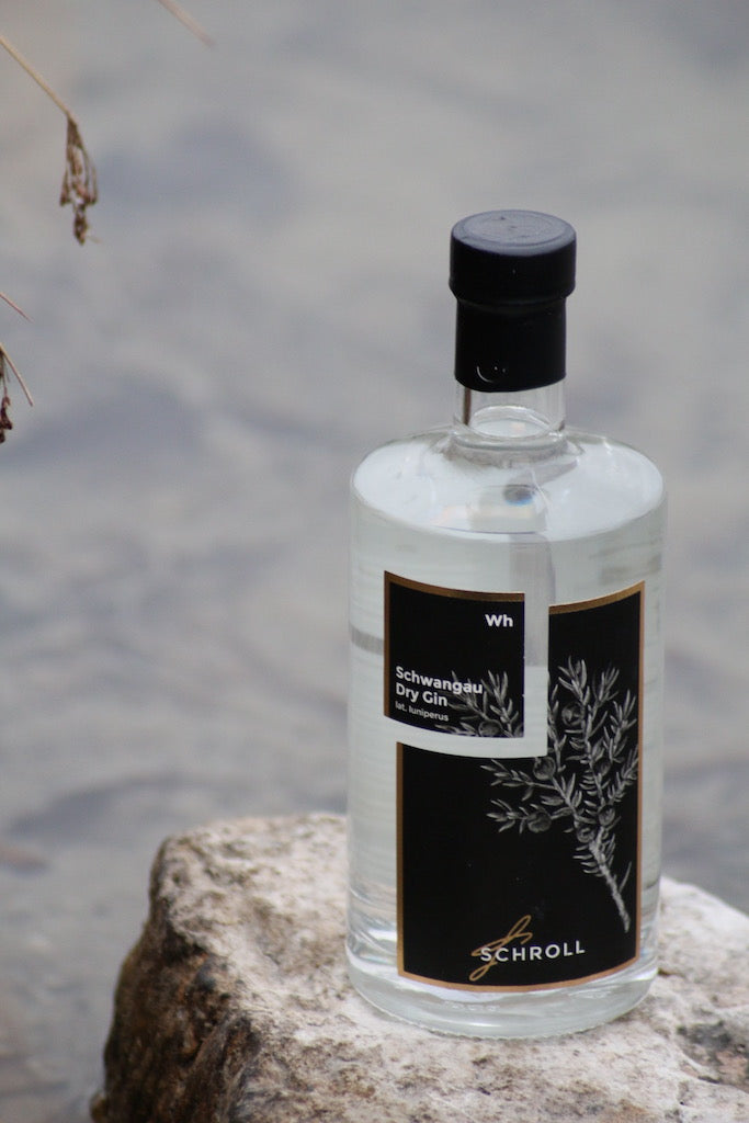 Schwangau Dry Gin Flasche mit schwarzem Etikett steht auf einem Stein am Ufer eines Gewässers.