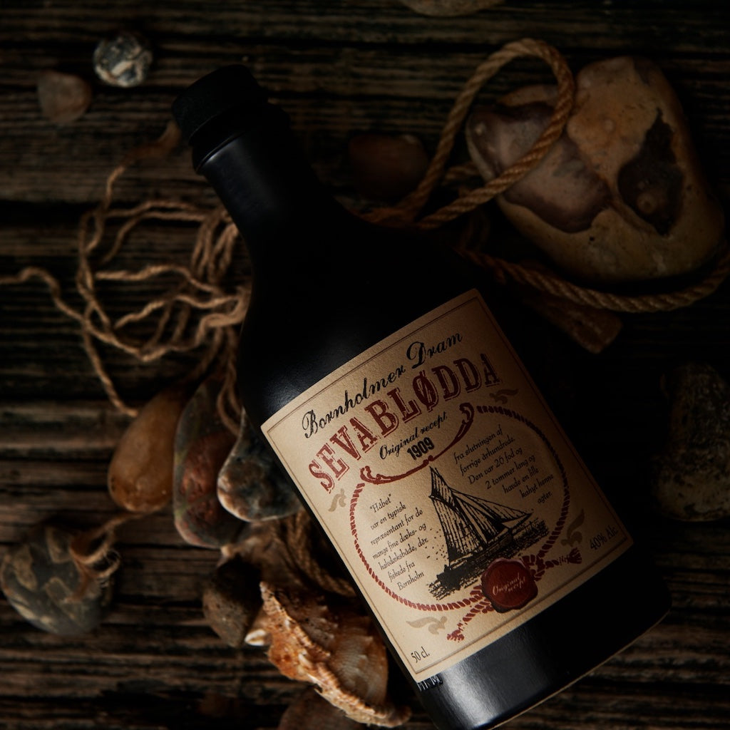 Bornholmer Dram Sevablødda 50cl, schwarze Flasche mit Vintage-Label, umgeben von Seilen und Steinen auf rustikalem Holzuntergrund.