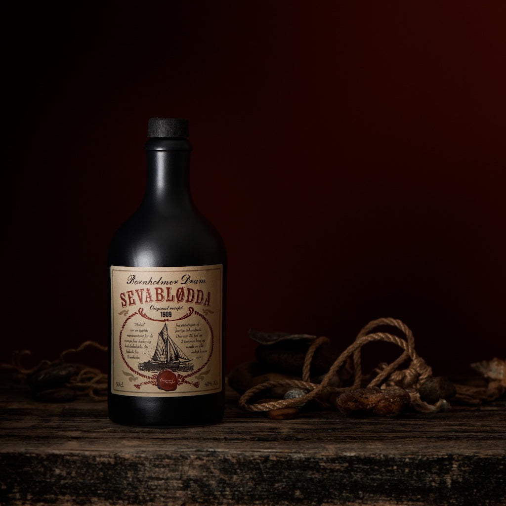 Bornholmer Dram Sevablødda 50cl, schwarze Flasche mit Vintage-Label, maritimes Design, rustikale Szene mit Seilen und Steinen.