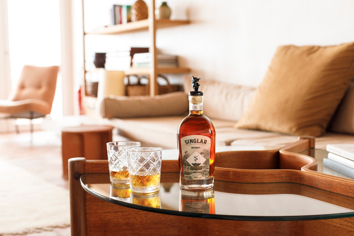 Flasche Singlar Whisky auf einem Glastisch, daneben zwei gefüllte Gläser, im Hintergrund ein Sofa mit Kissen.