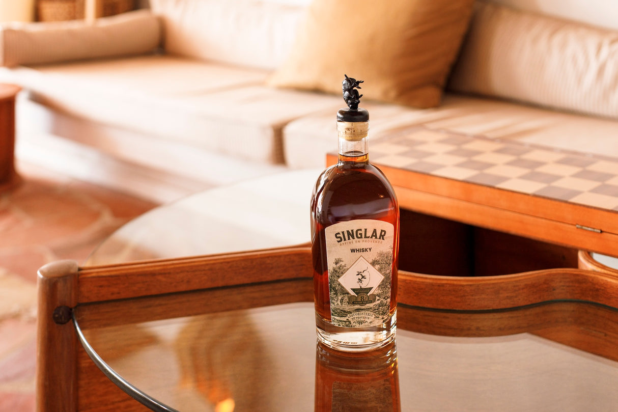 Flasche Singlar Whisky Blend 8Y mit Wildschweinverschluss auf einem Holztisch, im Hintergrund Sofa und Schachbrett.