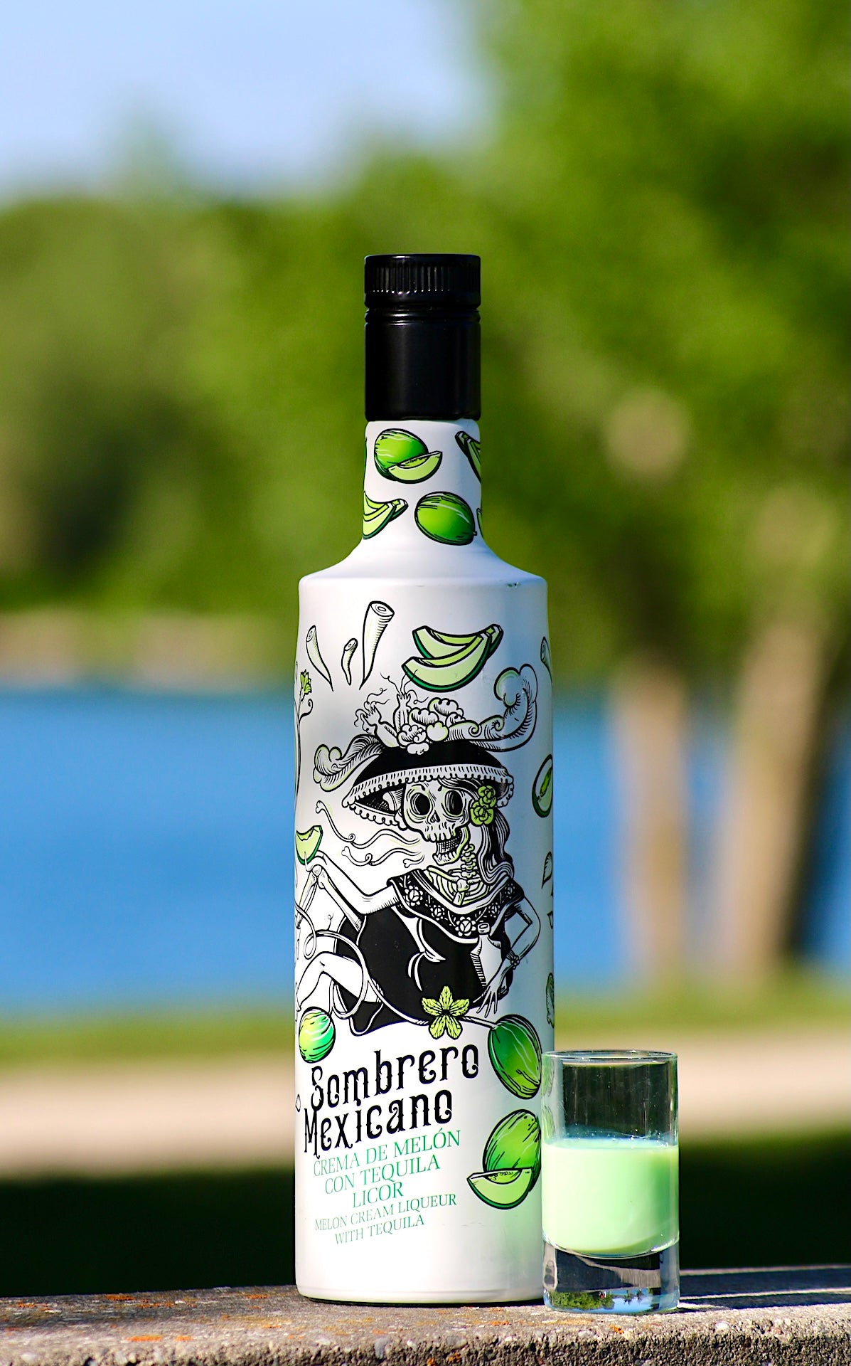 Flasche Sombrero Mexicano Melon Likör mit Tequila und grünem Shot im Glas, stehend vor See und grünem Hintergrund.