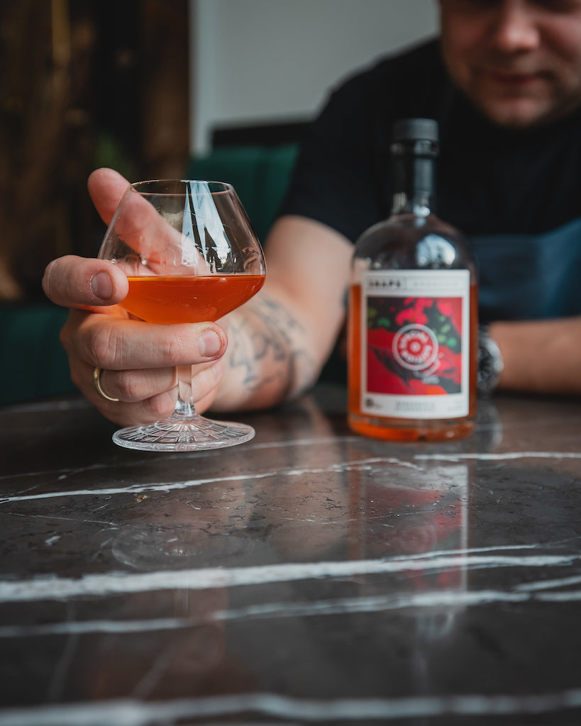 Hand hält ein Glas mit Bio-Schnaps Bornholm Rhabarber & Sternanis, im Hintergrund eine Flasche auf einem Marmortisch.