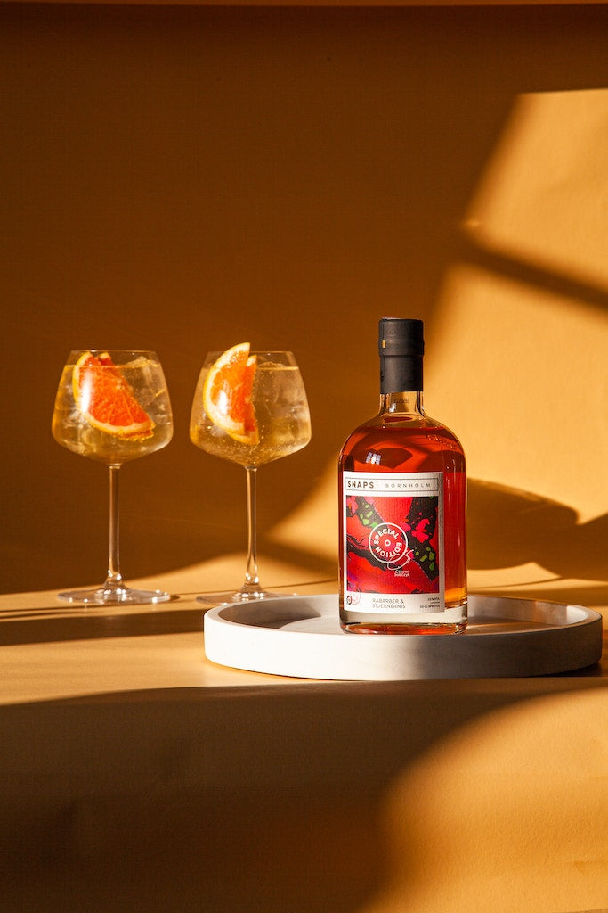 Bio-Schnaps Bornholm Rhabarber & Sternanis auf einem Tablett, daneben zwei Gläser mit Getränk und Orangenscheiben.