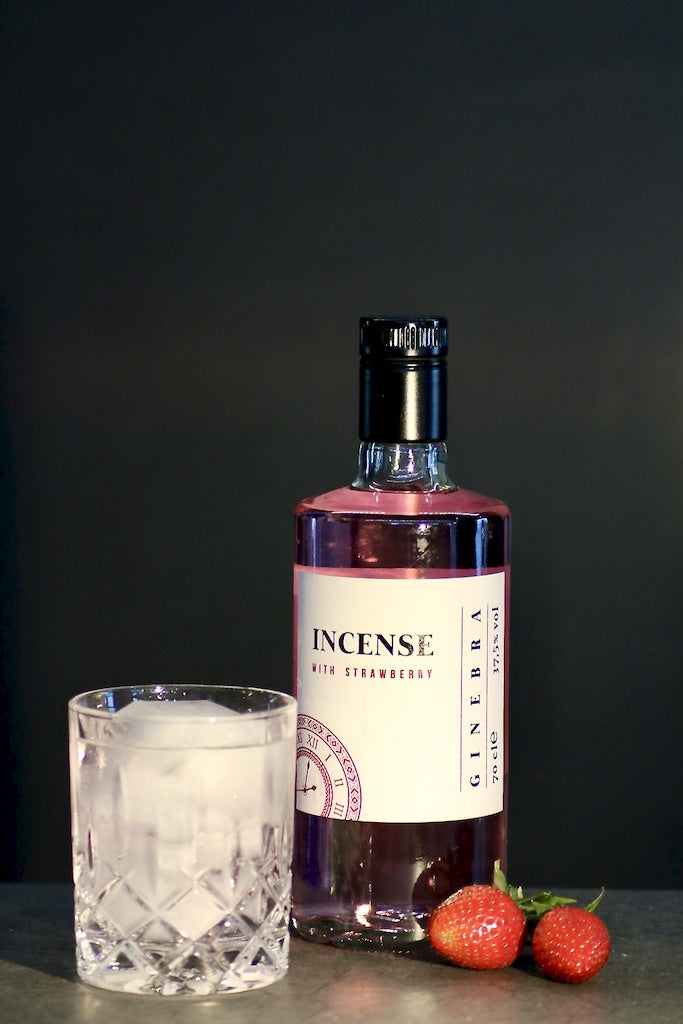 Flasche Incense Strawberry Gin mit Glas Tonic, Eiswürfeln und frischen Erdbeeren, präsentiert vor einem dunklen Hintergrund.
