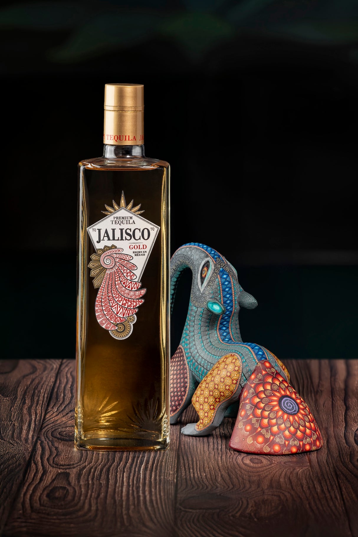 Tequila-Jalisco-Gold-Flasche auf Holzoberfläche neben einer bunt bemalten mexikanischen Alebrije-Figur.