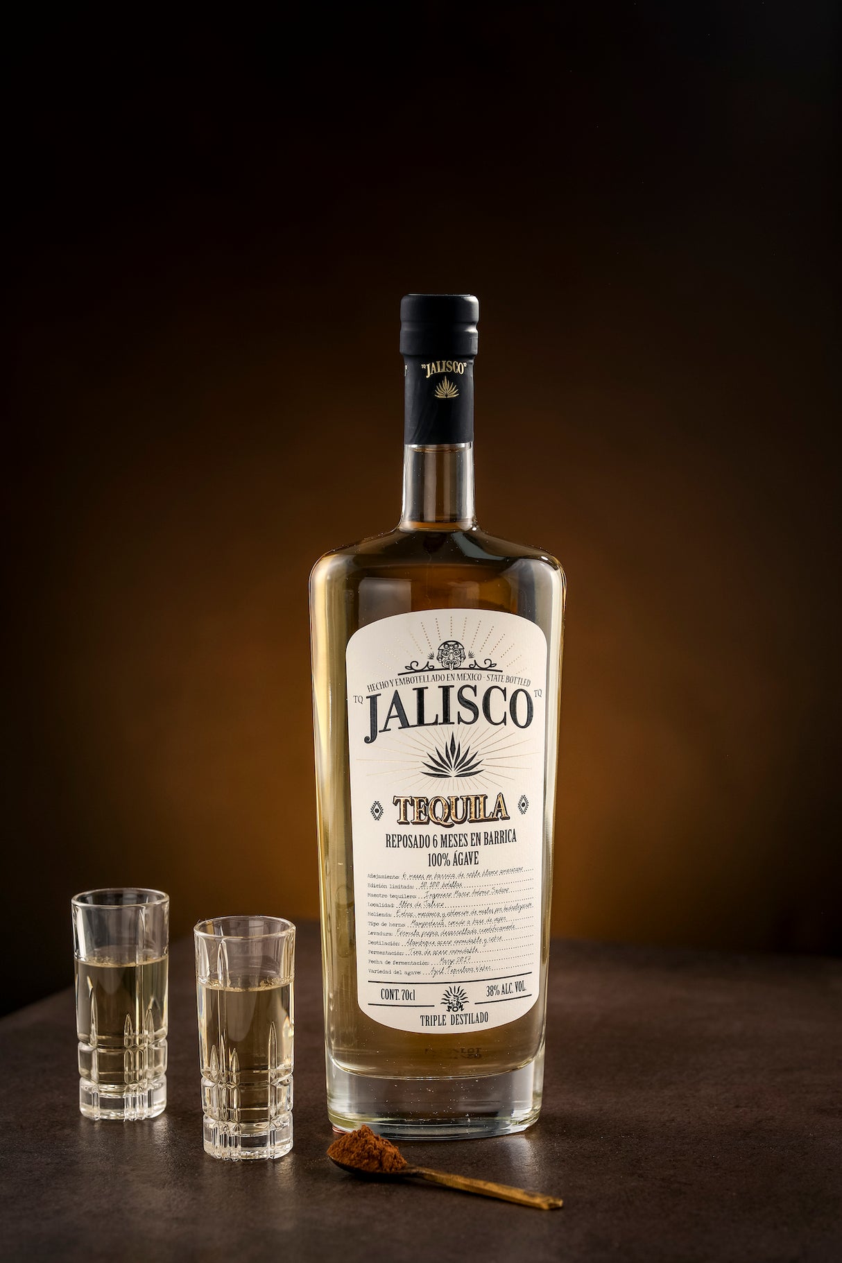 Tequila Jalisco Reposado Flasche mit zwei gefüllten Gläsern und einer Zimtstange auf einem dunklen Hintergrund.
