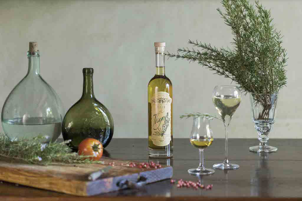 Flasche Fleur de Thym Likör auf Holztisch, Gläser mit Likör, Kräuter, Schneidebrett mit Tomate, Glasflaschen im Hintergrund.