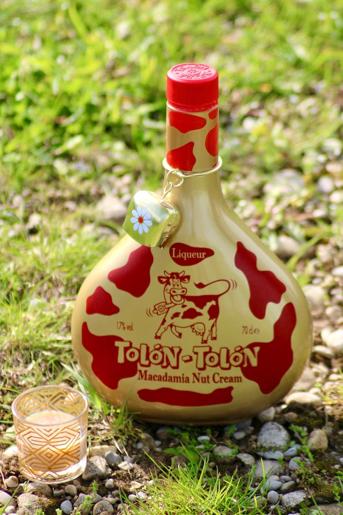 "Tolon-Tolon Macadamia Nut Cream Liqueur Flasche mit Glas, 70 cl, dekorative Kuhglocke, präsentiert auf Gras und Kies."