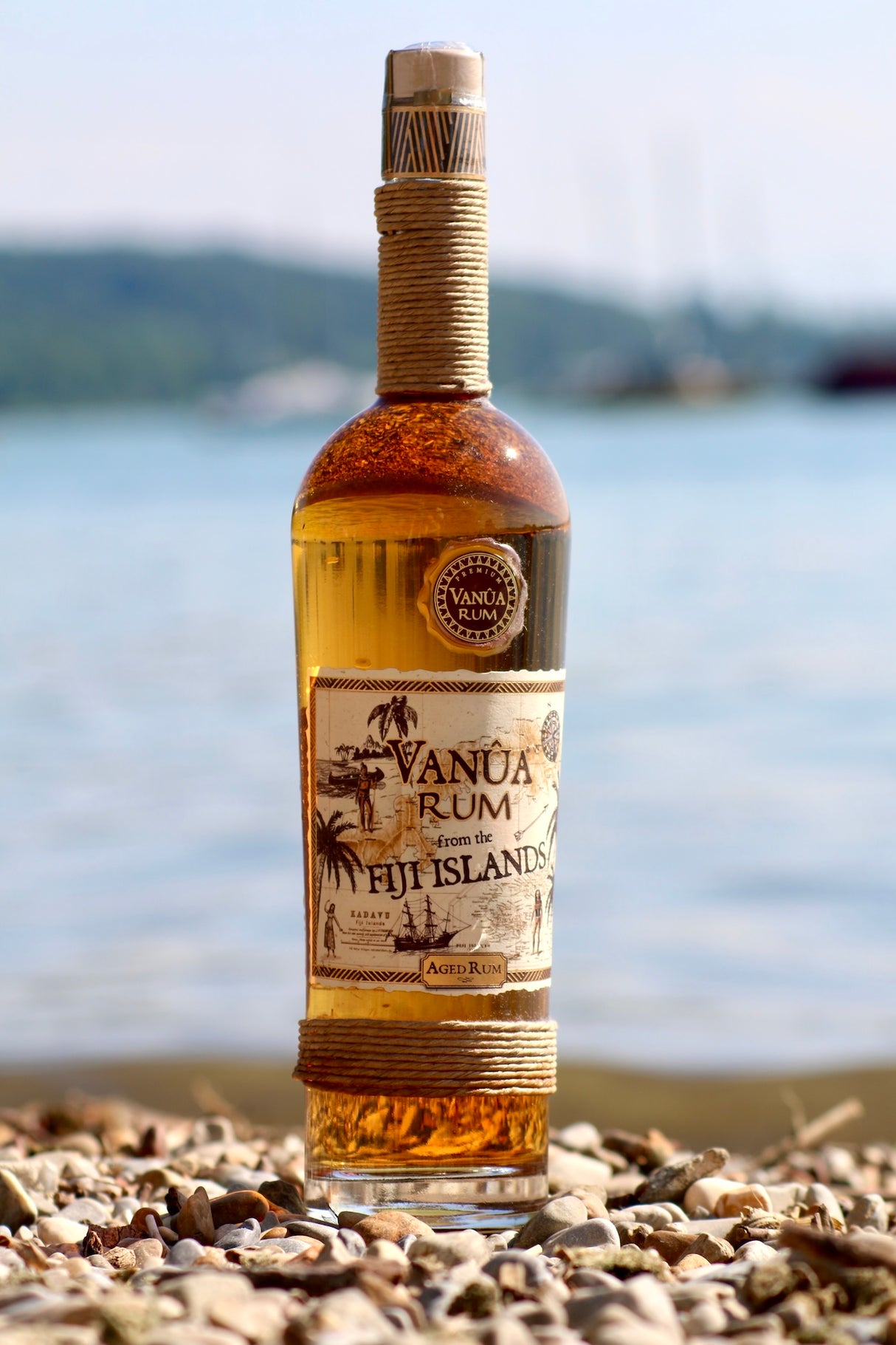 Vanua Rum Flasche von den Fidschi-Inseln auf Kieselstrand, im Hintergrund ruhiges Wasser und verschwommene Boote.