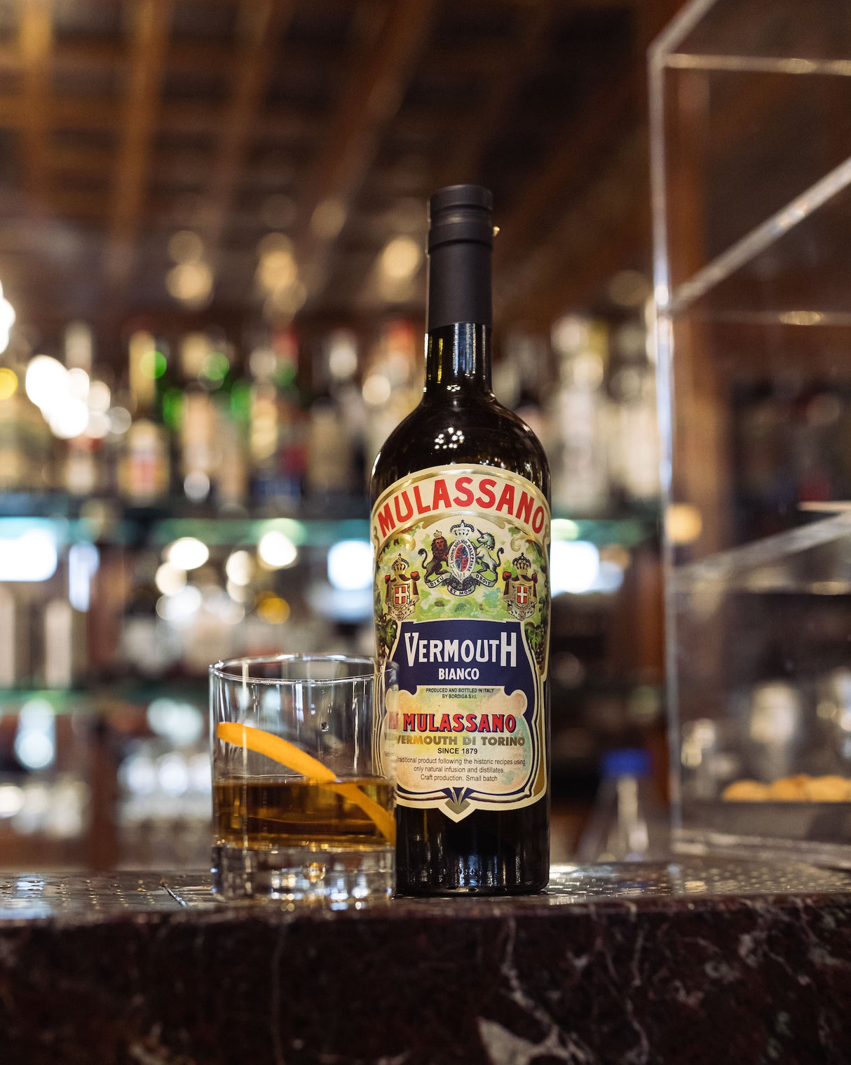 Vermouth Bianco Mulassano Flasche mit Glas auf Eis und Orangenzeste auf einer Bar, unscharfer Hintergrund