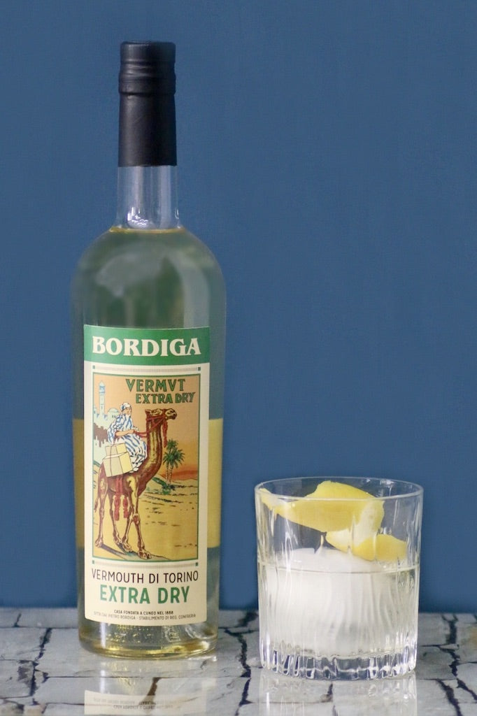 Flasche Bordiga Vermouth di Torino Extra Dry mit Kamelmotiv, neben Glas mit Eis und Zitronenschale vor blauem Hintergrund.