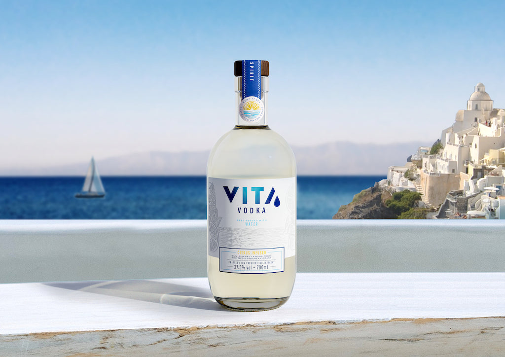 VITA Citrus Infused Vodka auf einer Terrasse mit Blick auf das Mittelmeer, ein Segelboot und weiße Häuser im Hintergrund.