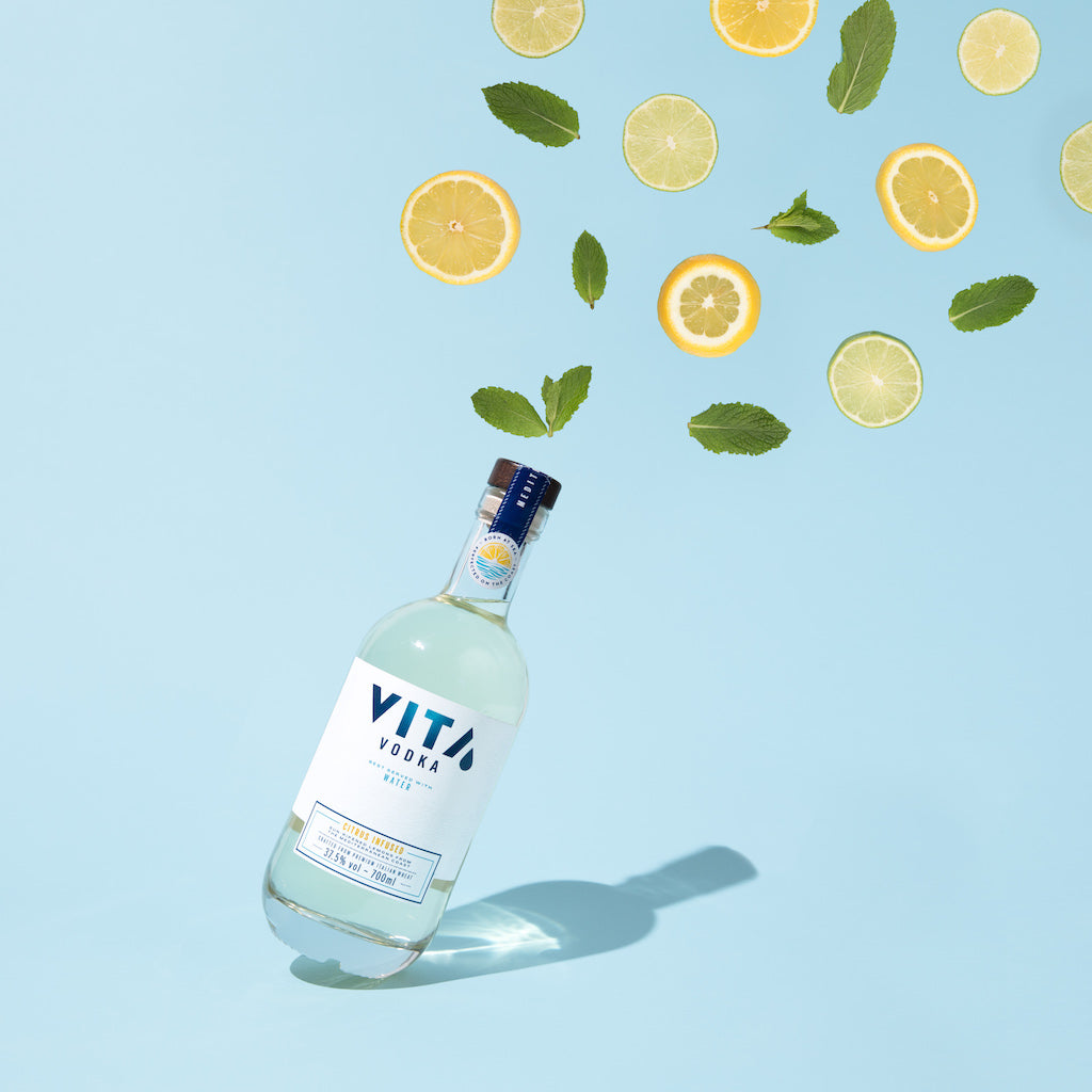 Flasche VITA Vodka auf blauem Hintergrund mit schwebenden Zitronen-, Limettenscheiben und Minzblättern.
