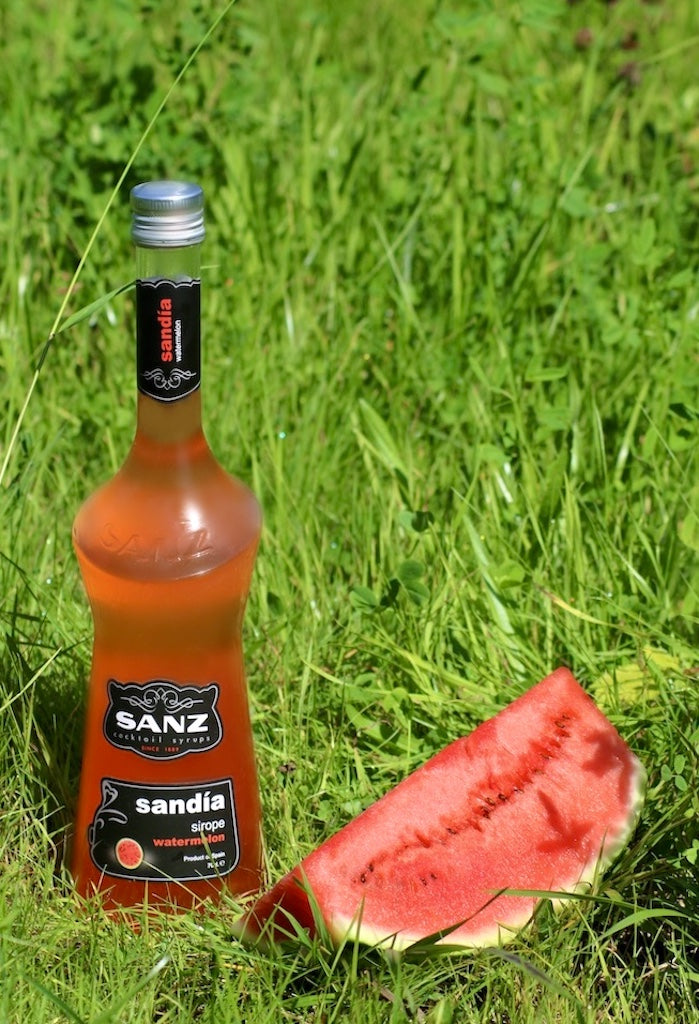 Wassermelonen-Sirup Sanz 0,7L in Glasflasche mit schwarzem Etikett, daneben ein Stück Wassermelone auf grünem Gras.