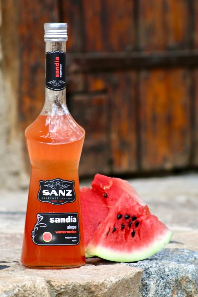 Wassermelonen-Sirup Sanz 0,7L in Glasflasche mit schwarzem Etikett, daneben frische Wassermelonenstücke auf Steinuntergrund.