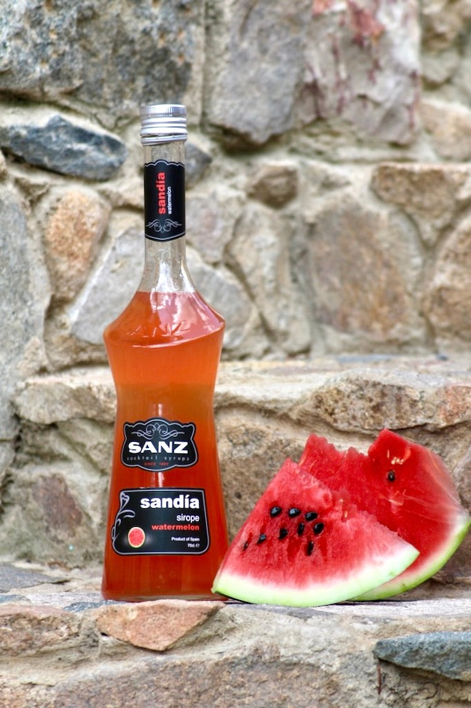 Wassermelonen-Sirup Sanz 0,7L in Glasflasche mit schwarzem Etikett, daneben frische Wassermelonenstücke vor rustikaler Steinwand.