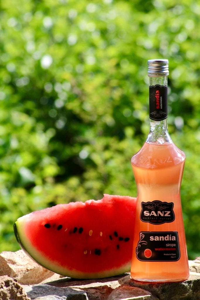 Wassermelonen-Sirup Sanz 0,7L in Glasflasche mit schwarzem Etikett, daneben ein Stück Wassermelone vor grünem Hintergrund.