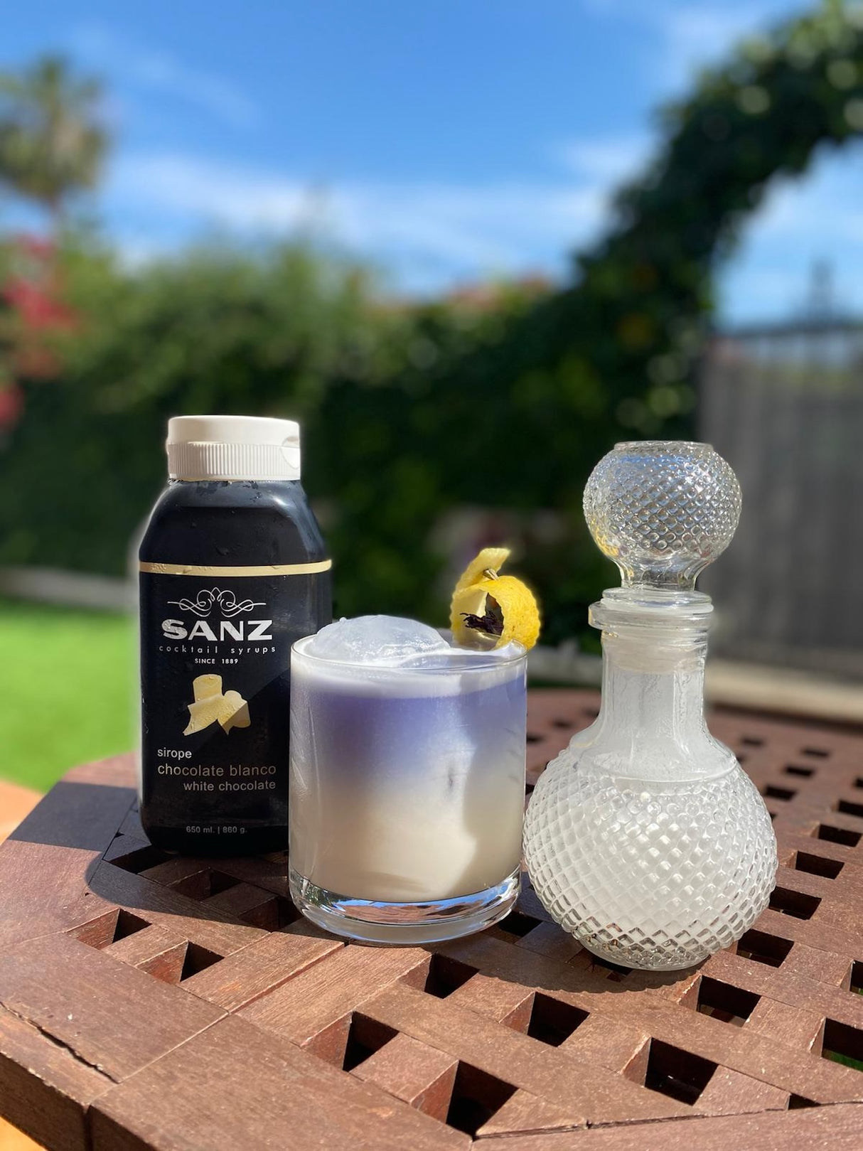 Sanz Sirup Weiße Schokolade mit Cocktail im Glas, dekoriert mit Zeste, auf Holztisch im Garten bei Sonnenschein.