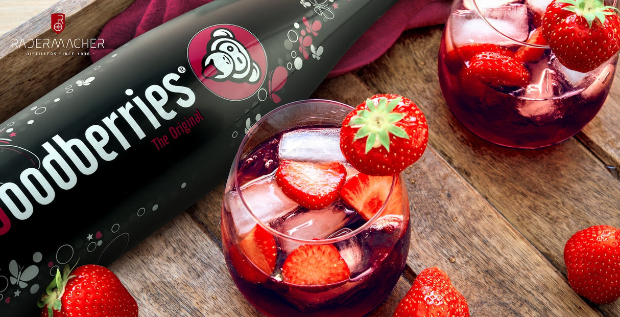 Woodberries-Flasche neben Cocktailgläsern mit Eiswürfeln und frischen Erdbeeren auf einem Holztisch.