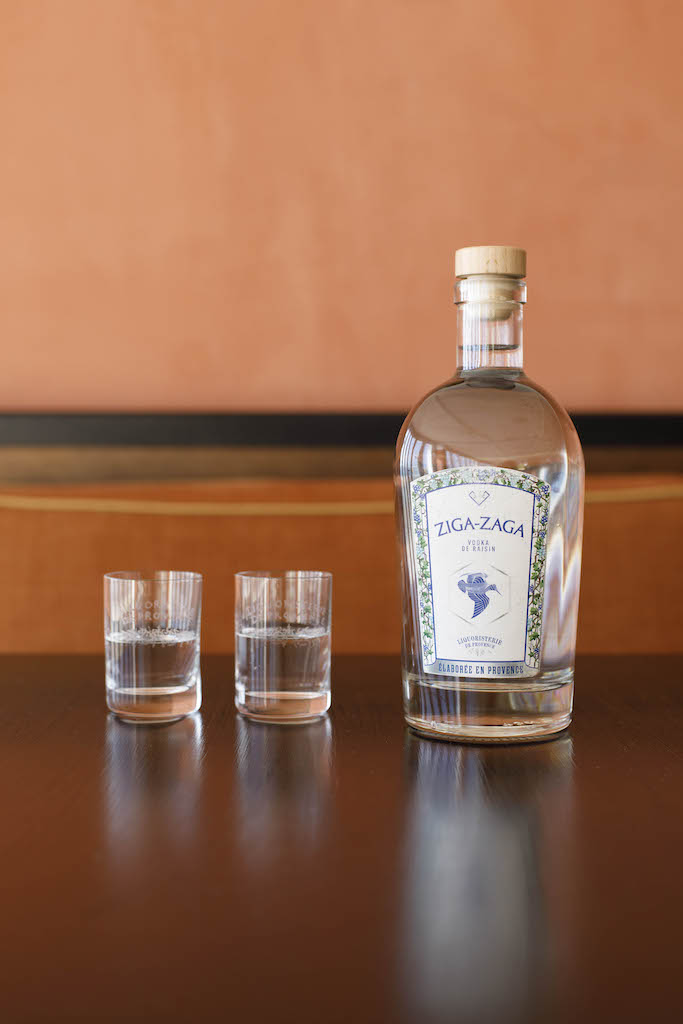 Flasche Ziga Zaga Vodka mit zwei gefüllten Gläsern auf dunklem Tisch vor orangefarbener Wand.