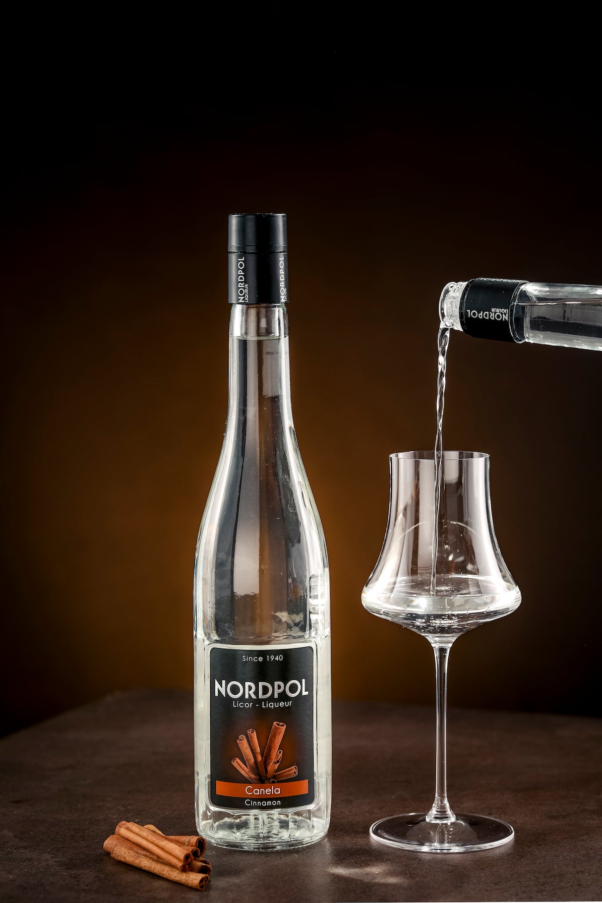 Flasche Nordpol Zimtlikör (Canela) neben Glas, in das Zimtlikör eingeschenkt wird, mit Zimtstangen im Vordergrund auf dunklem Hintergrund.