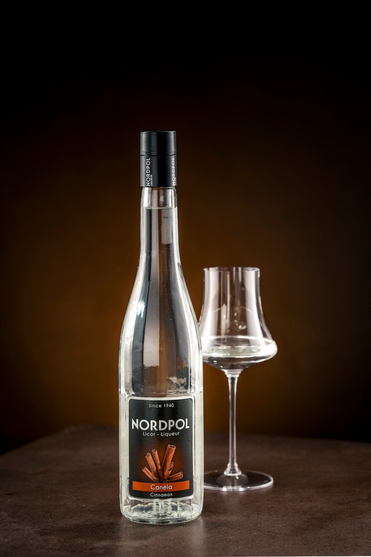 Flasche Nordpol Zimtlikör (Canela) neben leerem Glas, vor dunklem Hintergrund.