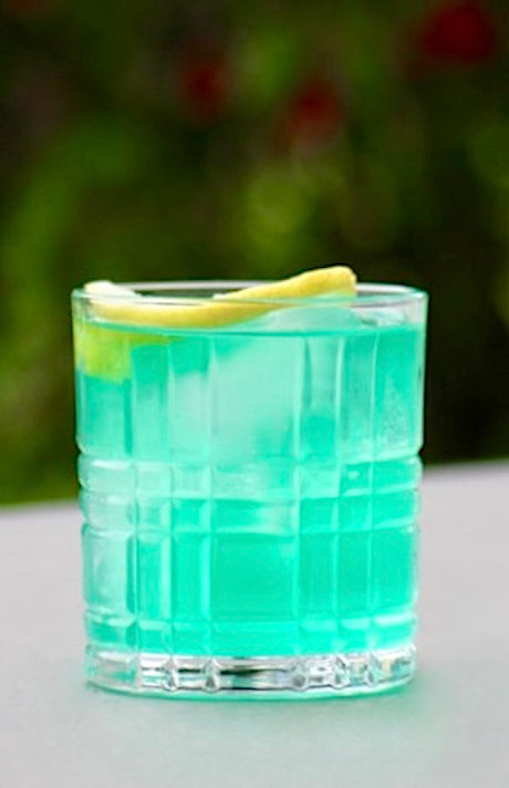 "Ein erfrischender Blue Lagoon Twist Cocktail in einem Whisky-Glas, serviert mit Eiswürfeln und einer Zitronenscheibe als Garnitur. Der Cocktail hat eine leuchtend blaue Farbe und vermittelt tropisches Flair.