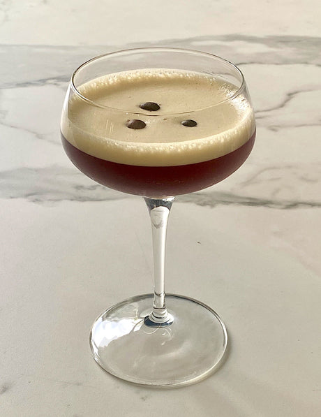 Grappa Espresso Martini in einer Coupette mit heller Schaumkrone und drei Kaffeebohnen auf der Oberfläche.
