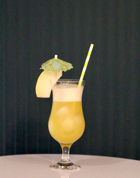 Melon del Sol Cocktail in einem Hurricane-Glas mit gelber Melonenscheibe, grünem Cocktail-Schirmchen und gestreiftem Strohhalm.