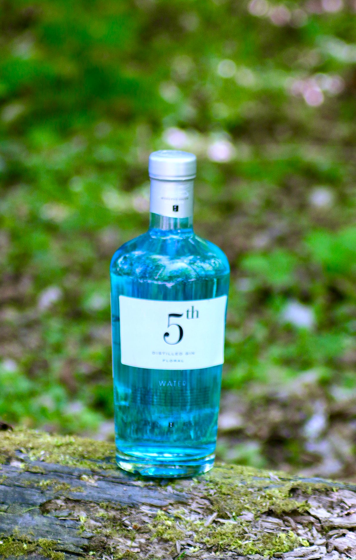 5th Gin Water blaue Flasche auf moosigem Baumstamm vor grünem Naturhintergrund
