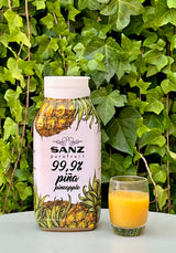 Sanz Ananaspüree 99,9 % mit Flasche und Glas mit gelbem Ananaspüree auf einem Tisch vor Efeu.
