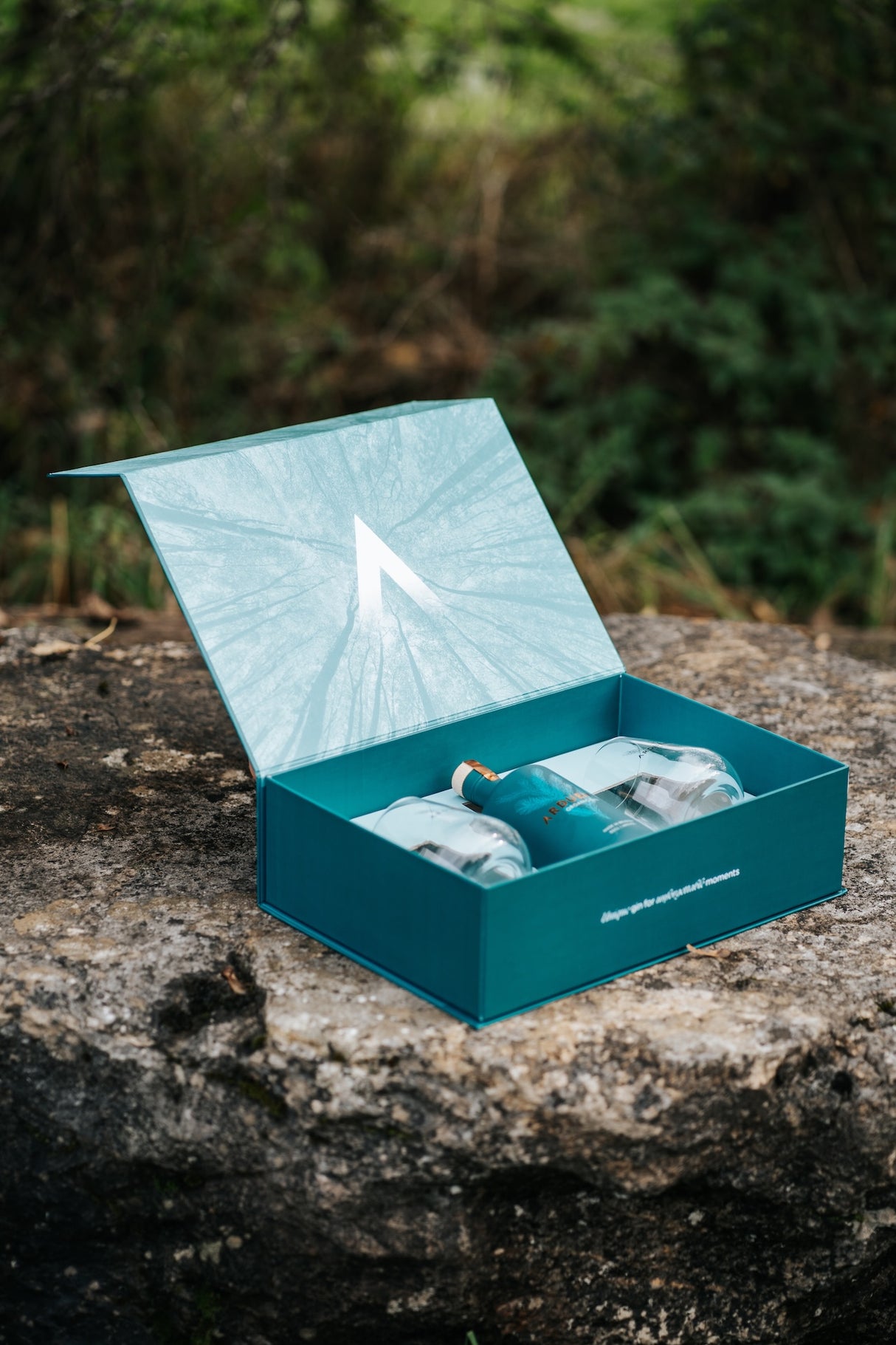 Geöffnete Arduenna Geschenkbox mit Bio Gin und zwei Gläsern auf einem Felsen vor grünem Hintergrund.