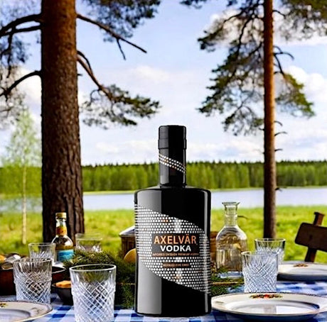 Axelvår Vodka auf einem gedeckten Tisch, umgeben von skandinavischer Landschaft, Bäumen und einem See im Hintergrund.