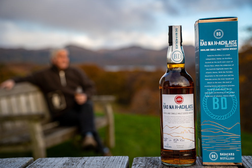 Flasche Bad na h-Achlaise Tuscan Oak Cask Strength Highland Single Malt Scotch Whisky mit Geschenkbox, im Hintergrund eine Person auf einer Bank vor einer malerischen Landschaft.