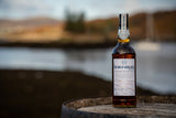 Flasche Bad na h-Achlaise Tuscan Oak Highland Single Malt Scotch Whisky auf einem Fass, im Hintergrund eine ruhige Landschaft mit Wasser und Bergen.