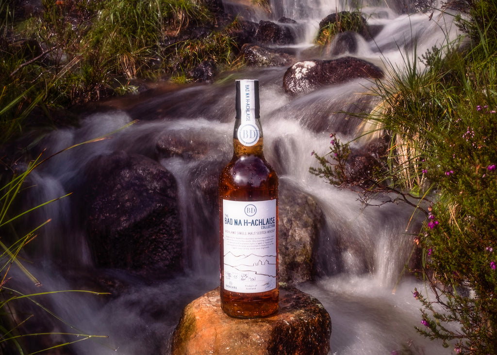 Flasche Bad na h-Achlaise Tuscan Oak Highland Single Malt Scotch Whisky auf einem Stein vor einem plätschernden Wasserfall in einer natürlichen Umgebung.