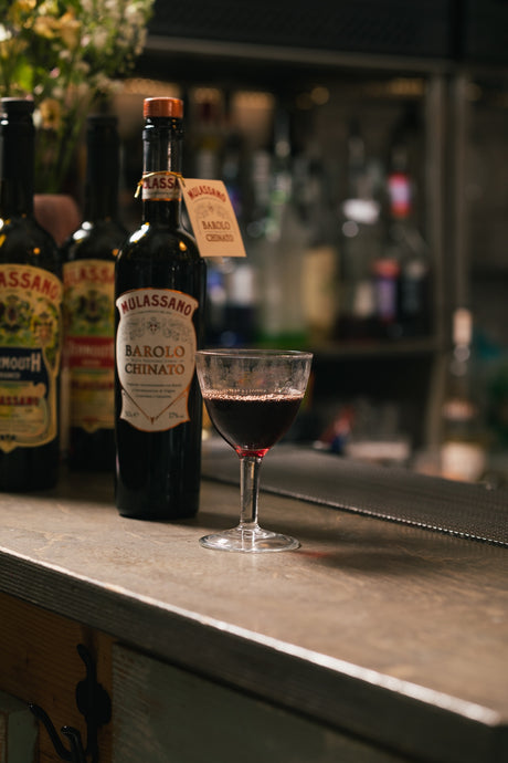 Mulassano Barolo Chinato Flasche mit gefülltem Glas auf Bartresen vor unscharfem Flaschenhintergrund