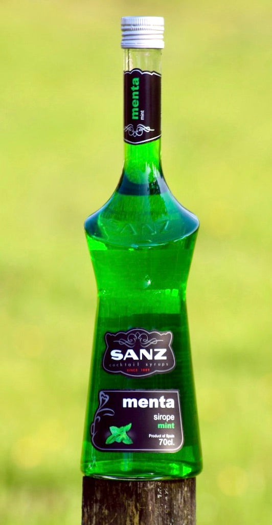 Grüne Flasche Sanz Minzsirup mit schwarzem Etikett und mintfarbenem Schriftzug, im Freien auf Holzstumpf platziert.