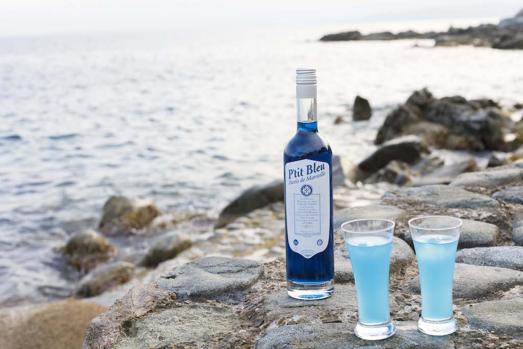 Flasche P'tit Bleu Pastis de Marseille und zwei Gläser mit blauem Getränk am Meer auf Felsen platziert.