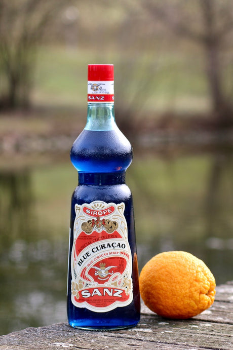 Flasche Sanz Blue Curacao Sirup mit rotem Etikett und Verschluss, 1 Liter, auf Holzfläche neben einer Orange.