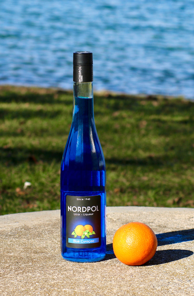 Flasche Nordpol Blue Curaçao Likör und eine Orange auf einem Stein, im Hintergrund Wiese und Wasser