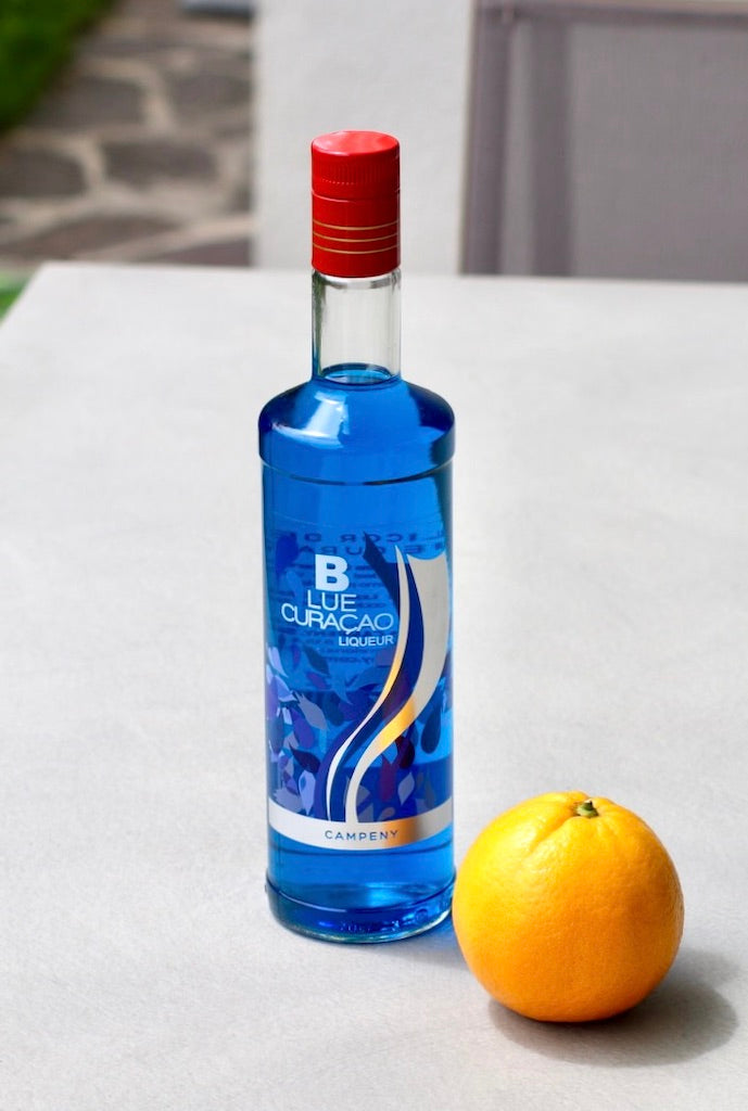 Flasche Campeny Blue Curaçao Likör neben einer Orange auf einer hellen Oberfläche. 