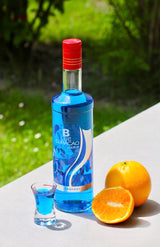 Flasche Campeny Blue Curaçao Likör neben einem Glas mit blauem Likör und einer aufgeschnittenen Orange, auf einer hellen Oberfläche, mit einem grünen Hintergrund.