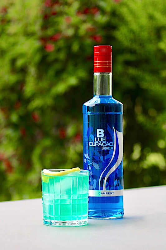 Eine Flasche Blue Curacao Likör von Campeny neben einem Blue Lagoon Twist Cocktail in einem Whisky-Glas, serviert mit Eiswürfeln und einer Zitronenscheibe, vor einem grünen Hintergrund.